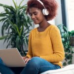 Shot of beautiful woman using her laptop while listening to music with headphones sitting on a puff at home.